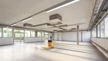 Large open commercial space with concrete floors, exposed ceiling, and overhead lighting. The space has large windows providing natural light and a forklift visible, indicating it may be used for storage or distribution purposes.