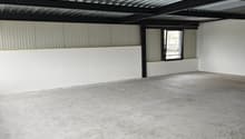 The image shows the interior of a warehouse or industrial space. The ceiling is made of corrugated metal panels, and there are black metal beams supporting the structure. The floor is a plain concrete surface. The space appears to be empty and unfinished,