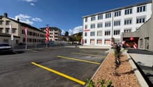 A multi-story commercial building with a large parking lot in front. The building has a modern, white facade with numerous windows. The parking lot is marked with yellow lines and contains several parked cars. There are some decorative plants and flowers