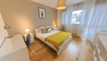 Spacious bedroom with hardwood flooring, white walls, large windows, a double bed with yellow bedding, nightstands, and a pendant light fixture.