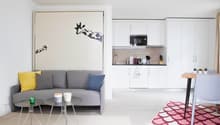 white living room with white walls, gray couch with pillows, white cabinets, wooden microwave, coffee machine, white coffee cups, carpet, white dining table with white chair, pink rug, white table with white cups
