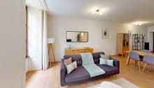Spacious living room with hardwood floors, white walls, large windows, gray sofa, wooden TV stand, and shelving unit. The room also features a balcony or terrace.