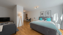 Bedroom with wooden floor, white walls, bed with blue pillows, two nightstands, bedside lamps, wall-mounted pictures, a table and chairs in a corner, a TV, a lamp, and a chair.