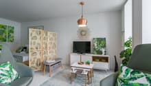 Bright and airy living room with white walls, hardwood floors, and tropical-inspired decor. Features a copper pendant light, a wooden screen divider, a TV stand with storage, and a seating area with a gray sofa and armchair. The room also has potted plant