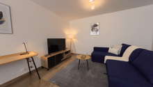 Living room with a blue sofa, wooden desk, TV, lamp, and rug