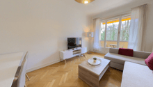 Spacious living room with large windows providing a view, hardwood flooring in a herringbone pattern, white walls, a TV stand, a sofa, and a coffee table with decorative items.