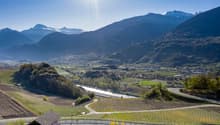 The image shows a picturesque mountain landscape with snow-capped peaks in the background. In the foreground, there are rolling hills covered in vineyards and farmland, with a small town nestled in the valley below. The scene is bathed in warm, golden sun