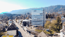 Office building with many windows, surrounded by other buildings, trees, and mountains in the background