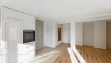 Open-concept living space with white walls, hardwood floors, built-in kitchen with oven and dishwasher, and a hallway leading to other rooms