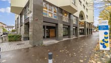 A multi-story commercial building with a modern, glass-fronted design. The building has a stone facade and a covered entrance. There are several businesses operating in the building, including Kita and Insign. The surrounding area has trees with autumn fo