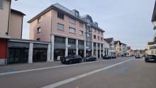 A street with a few parked cars in front of buildings, some with balconies and shops