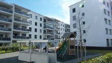 Multi-story apartment building with balconies, parking area, and a children's playground in the foreground.