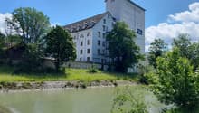 Large white building with multiple windows, situated next to a river, surrounded by trees and grass.