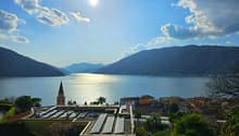 The image shows a scenic view of a lake surrounded by mountains. The lake is reflecting the sunlight, creating a shimmering effect on the water. In the foreground, there is a town with colorful buildings and a church steeple visible. The landscape is lush