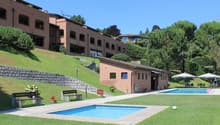 large brick building on the hill, large grassy yard, small pool, small house, benches, patio chairs and tables, large umbrellas
