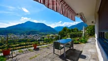 Balcony with a stunning view of the mountains and valley below. The balcony has a striped awning, a table and chairs, and potted plants. The surrounding landscape is lush and green, with mountains in the distance under a blue sky with clouds.