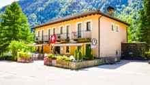 2-story building with a yellow exterior, balconies, and a Swiss flag. The building is surrounded by trees and has a patio area in front with tables and chairs. The building appears to be a hotel or restaurant located in a mountainous, forested area.