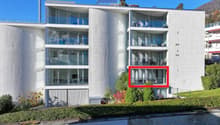Modern multi-story apartment building with glass balconies and terraces, surrounded by landscaping and a water feature.