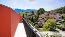 The image shows a balcony or terrace with a view of a small town surrounded by mountains. The buildings in the town appear to be a mix of residential and commercial structures, with trees and greenery visible throughout. The balcony has a red railing and