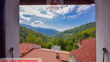 View of a mountainous, forested landscape from a window or balcony. The image shows red-tiled roofs of houses in the foreground, with a backdrop of lush green hills and mountains under a blue sky with clouds.