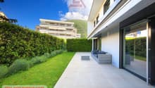 A white building with glass windows, tiled patio leading to a sofa on a terrace, green hedges and grass lawn, and a mountain in the background.