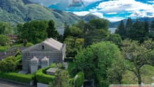 A two-story villa with a view of the mountains and lake, large landscaped garden, and a nearby road.