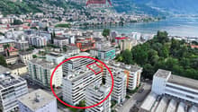 Aerial view of a residential building complex with multiple buildings, surrounded by trees and a lake, mountains in the background.