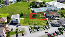 The image shows an aerial view of a residential area surrounded by vineyards and greenery. There is a parking lot visible in the foreground, and several buildings and houses can be seen in the background. The area appears to be a small town or village nes