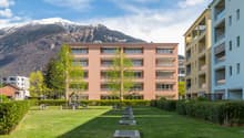 Two story apartment building, white and pink exterior, several balconies, green lawn, bushes, playground, trees, mountains in the background