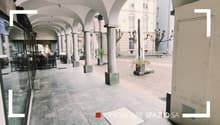 The image shows an outdoor area with arched walkways, columns, and potted plants. The ground is paved with stone tiles, and the surrounding buildings have a historic architectural style. This appears to be a commercial or public space, potentially a plaza