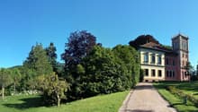 Large historic mansion with a tower, surrounded by lush greenery and a well-manicured garden with a paved walkway leading to the entrance.