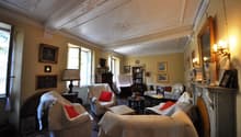 Spacious living room with ornate ceiling, large windows, antique furniture, artwork, and a fireplace