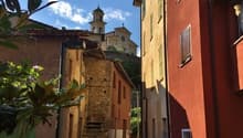 Narrow alleyway with colorful buildings, stone walls, and a church steeple visible in the background. The scene has a charming, historic European feel with lush greenery and a bright, sunny sky.