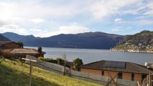 The image shows a scenic view of a lakeside town surrounded by mountains. The town has a mix of residential and commercial buildings with red-tiled roofs. The lake is visible in the foreground, with mountains rising in the background. The landscape appear