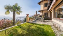 Two-story Mediterranean-style house with a terrace, palm tree, and ornate wrought-iron railing overlooking a grassy yard and mountains in the background. The house has a stone exterior, red tile roof, and decorative architectural details.