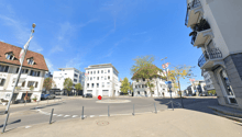 The image shows a street view of a city center. There are various buildings, including office buildings and shops, lining the street. The street has a roundabout in the center, and there are parked cars and pedestrians visible. The buildings have a mix of