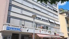 A building with several floors, exterior paint in pink and white, entrance with door and window, ground floor shops with signs, awnings, and signage, street light and parked motorbike