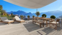 Large wooden deck with outdoor furniture, including lounge chairs and a dining table, with a panoramic view of the mountains and lake in the background. The deck is covered by a canopy and surrounded by potted plants and trees.