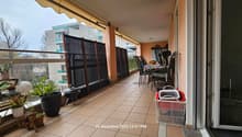 A balcony with various potted plants, a dining table with chairs, a storage box, a fan, a grill, and a rug. The balcony offers a view of the street and the surrounding area, with buildings and trees visible.
