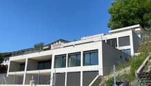 A modern, multi-story building with a balcony or terrace visible, situated on a hillside with lush greenery surrounding it. The building has a sleek, contemporary design with large windows and a gray exterior.