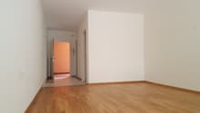 A hallway with wooden floors and white walls, leading to rooms with closed doors and a visible radiator. There's a white sheet on the floor at the end of the hallway.