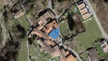 The building is an old medieval-style stone house located in the village. The surrounding area is rural, with houses, farmland, and some trees. A blue arrow points to the main entrance of the building.