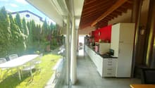 Spacious kitchen with large windows overlooking a garden, wooden ceiling beams, white cabinets and drawers, red accent wall, dining table and chairs