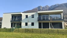 Two-story apartment building with a modern, minimalist design. The building has large windows, balconies, and a garage. It is surrounded by a grassy lawn and has a scenic mountain backdrop.