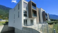 Modern multi-story apartment building with white and dark gray exterior, large windows, and balconies. The building is situated on a raised ground floor with a concrete retaining wall and fencing around the property.
