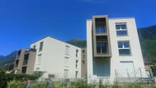 Multi-story apartment building with modern, minimalist design featuring large windows, balconies, and a grassy area with playground equipment in the foreground. The building is set against a backdrop of mountains under a clear blue sky.