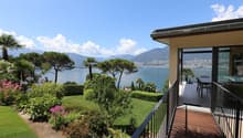 Luxury villa with a panoramic view of a lake surrounded by mountains. The property has a large balcony or terrace overlooking a well-manicured garden with palm trees, shrubs, and colorful flowers. The villa appears to be situated on a hillside, providing
