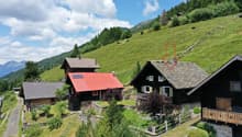 The image shows a picturesque mountain village nestled in a lush, green landscape. The village consists of traditional Swiss-style chalets with wooden facades and slate roofs. There are several buildings with balconies or terraces visible. The village is