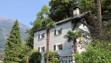 White building, brown shutters, steep slope roof, surrounded by trees, wooden fence, red pole, mountain in background