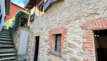The image shows the exterior of an old stone building with wooden beams and shuttered windows. The building has a stone facade with some brick accents, and there is a set of stairs leading up to the entrance. The surrounding area appears to be a quaint, h